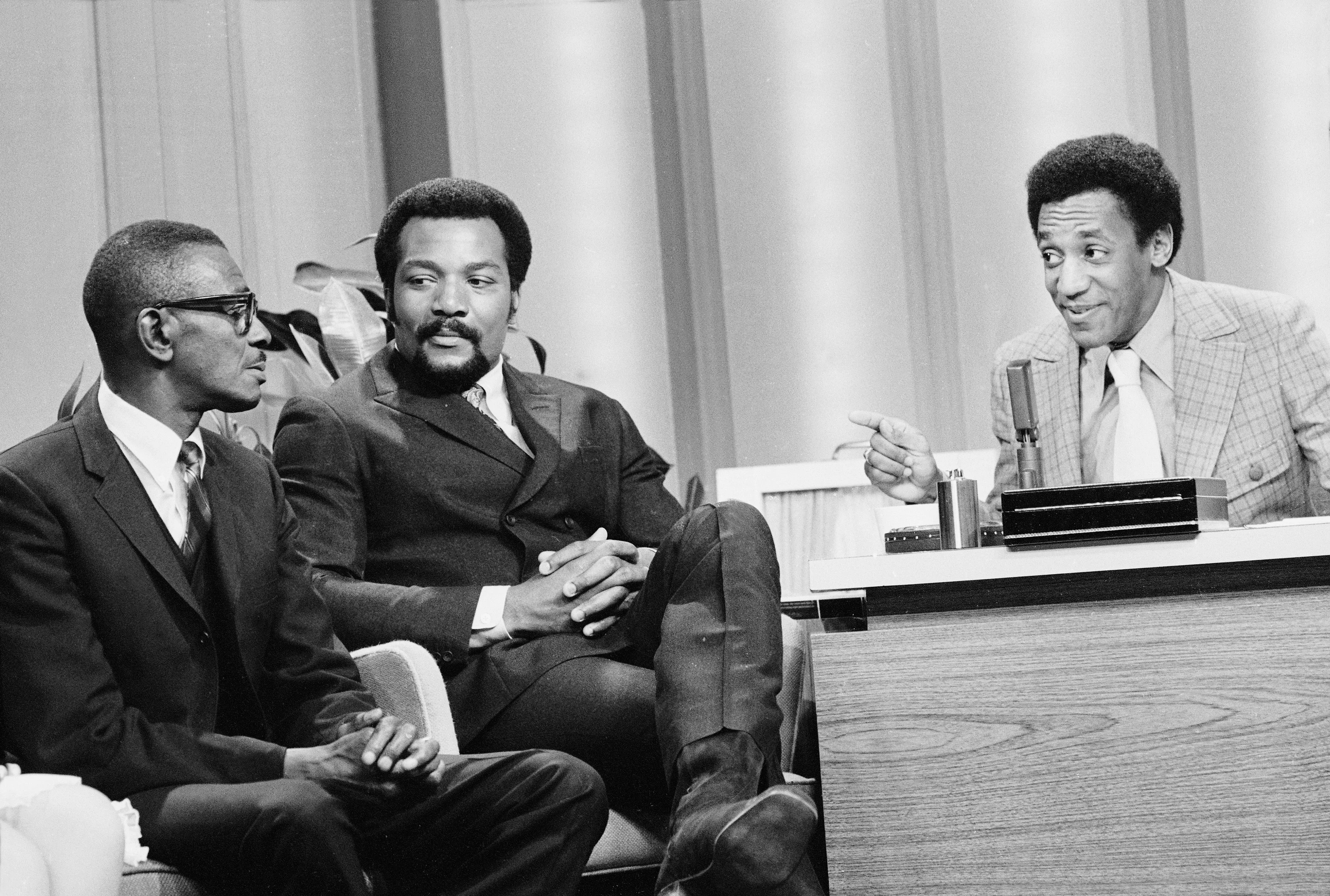 Bill Cosby, filling in as guest host on  The Tonight Show,  interviews Satchel Paige as football legend Jim Brown looks on, amused.