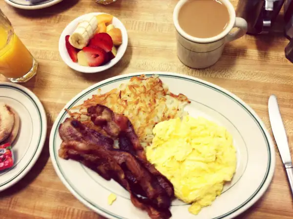 Bacon on plate with scrambled eggs and hash browns, with coffee and fruit salad and orange juice