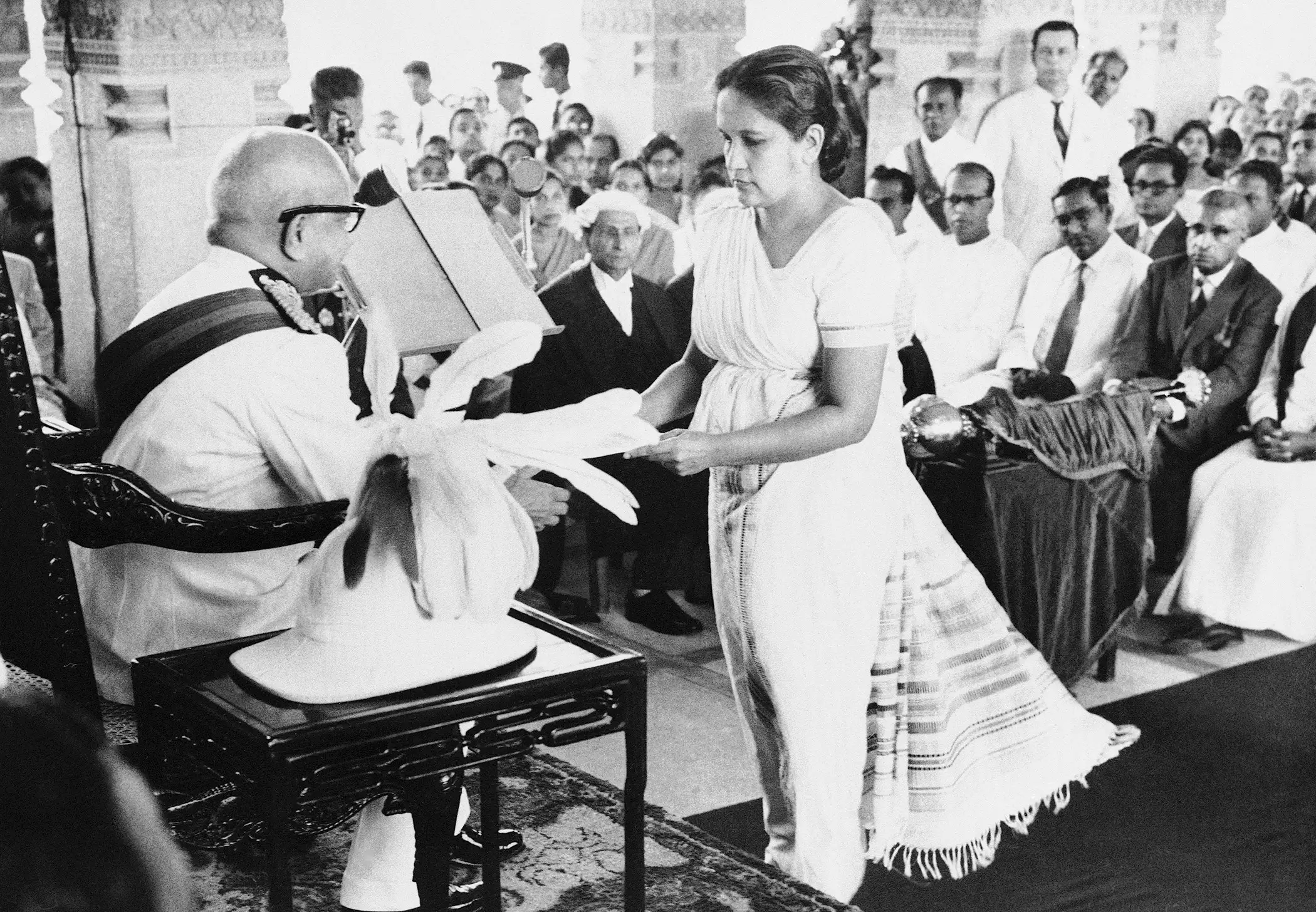 The Prime Minister of Ceylon, Mrs. Sirimavo Bandaranaike, hands over a copy of the throne speech to Sir Oliver Goonetilleke, the Governor General of Ceylon, at the ceremonial inauguration of the New Ceylon Parliament in Colombo on August 12, 1960. In the centre background wearing a wig and gown, is Ralph Deraniyagala clerk of the Ceylon House of Representatives. At right are (left to right) T.B. Ilangaratne the minister of trade: Felix Dias Bandaranaike minister of finance: and Dudley Senanayake, leader of the opposition (wearing a dark suit).