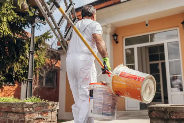 A man carrying a ladder and paint walking towards a house