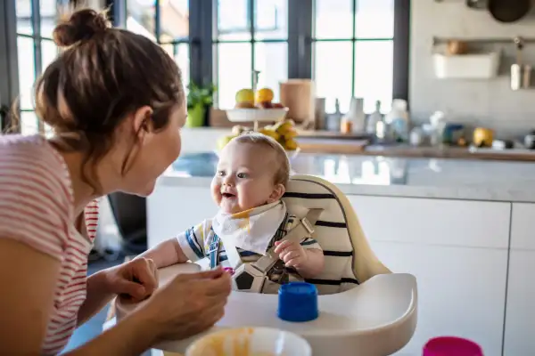 Mother feeds her baby son in high chair at home