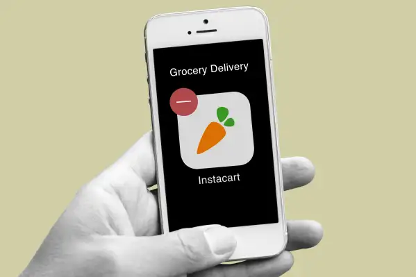 A closeup of a hand with a smartphone showing the Instacart app icon with a delete sign on the top left corner