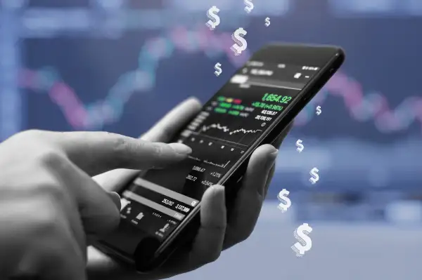 A close up of a hand holding a phone purchasing stocks on a trading app with some dollar signs in the background.