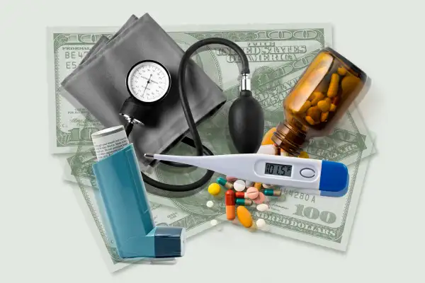 A bottle of pills, an inhaler, a thermometer and a blood pressure cuff with money in the background