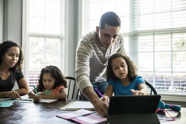 Mother And Father Helping Their Two Children With Homework At Kitchen Table