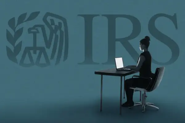 A woman sits on a desk working on her laptop while the IRS logo is in the background