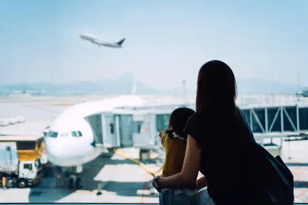 Young Asian mother embracing her cute little daughter looking at airplane through window at the airport while waiting for departure