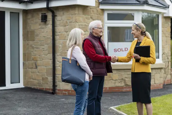 A real estate agent shaking hands with a senior man outside a bungalow that he has just bought with his wife, who is standing next to him