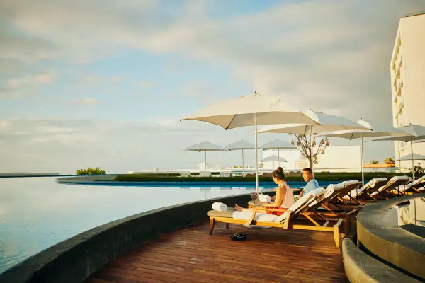 Wide shot of woman working on laptop while sitting in lounge chair with husband by pool at tropical resort