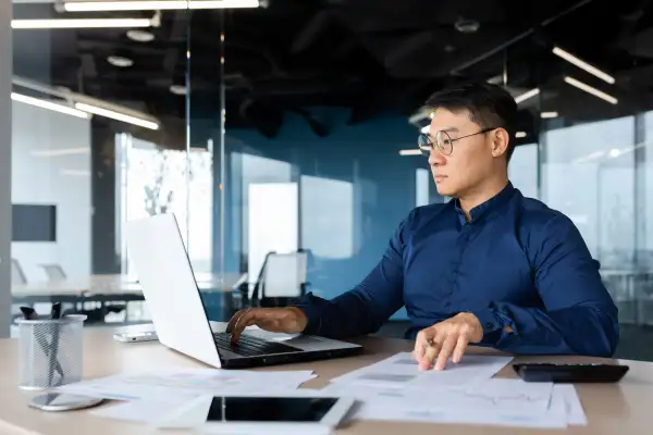 Man using a laptop in an office looking over some documents at the same time