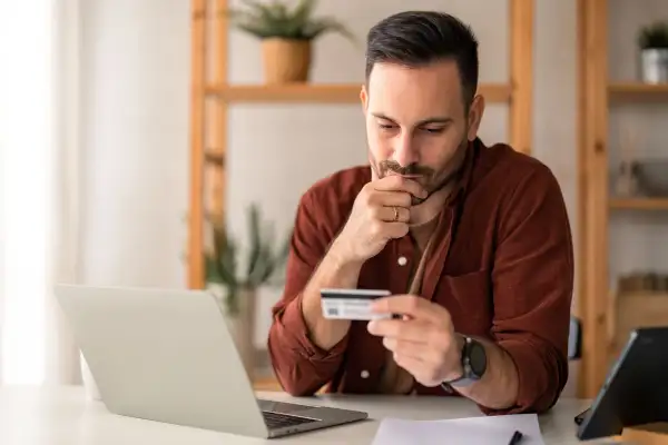 Man holding a credit card looking concerned sitting at a desk with an open laptop