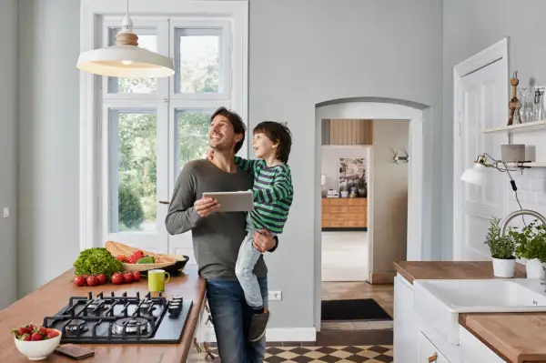 Father holding his son in his arms, in a beautiful kitchen, holding a tablet and looking at his light fixture